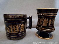 Two black decorative cups, one handled mug and one footed cup, with 24K gold Greek classical figure designs and Greek key patterns.