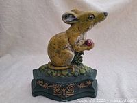 Front view of cast iron mouse door stop showing mouse in sitting position holding a red object, with decorative dark green and gold painted base.