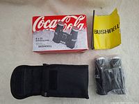 Photo showing the binoculars with Coca-Cola branded caps on eyepieces, original Coca-Cola box, yellow Bushnell pamphlet, and black carrying case.