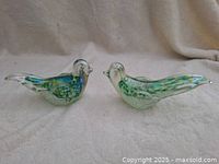 Pair of Murano style glass birds displayed side by side on a neutral fabric background, highlighting their iridescent coloration and shape.