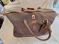 Top view showing brown textured fabric duffle bag with leather zipper pull, handle, and luggage tag with logo patch.