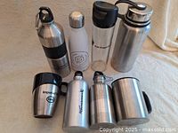 Photo showing 6 assorted water bottles and 2 stainless steel mugs arranged on a beige surface.