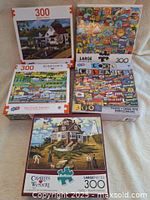 Set of five 300-piece puzzles stacked, showing box art featuring various scenes including coastal house, collages, and townscapes.