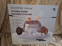Front of the box showing the Sharper Image S'mores Maker with flameless roaster and serving tray holding marshmallows, chocolate, and graham crackers.
