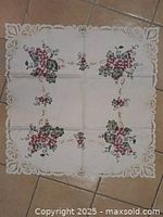 Square fabric table cloth with clusters of pink and purple embroidered flowers and scalloped lace trimmed edges laid flat on tiled floor