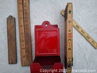 Photo showing three vintage folding wooden measuring rulers in different lengths alongside a red tin match holder with embossed design, all items laid flat on a white fabric background.