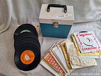 Image shows over 60 vintage 45 RPM vinyl records with various labels including Columbia and Playhouse, some in original colorful paper sleeves, displayed alongside a blue and white hard carry case with black handle.