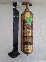 Vintage brass fire extinguisher and black metal wall mount shown from front view, label visible