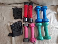 Photo showing four sets of dumbbells in red, blue, green vinyl coatings and gray metal hex design, alongside one pair of black fingerless training gloves with padded palms and velcro straps.