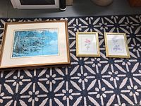 Three framed artworks displayed on a patterned carpet, includes a large winter scene and two smaller floral prints.