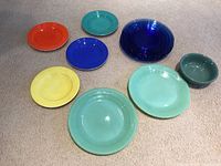 Top view showing 11 total pieces: 5 blue Depression glass plates, 2 green Jadeite Fire King plates, 4 assorted colored plates in different colors, and 1 green stoneware bowl.