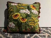 Front view of square pillow with colorful floral embroidery and brown crocheted trim on a carpeted surface.