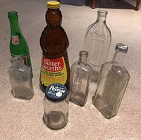 Front view of seven various vintage bottles on carpet including Mrs. Butterworth's syrup bottle, green 7UP bottle, clear bottles embossed with text and a bottle with metal lid.