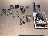 Photo showing all kitchen utensils laid out including hand crank can openers, ladle, peeler, masher, egg beater, grater and other tools.