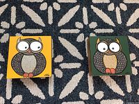 Two owl paintings side by side on a patterned rug, one with yellow background and one with green background.