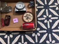 Overview of random items in a cardboard box on patterned carpet showing clocks, thermometer, knife with sheath, and empty soda can