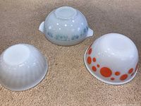 Three vintage milk glass bowls shown upside down to reveal brand marks and design patterns: Pyrex Butterprint Cinderella bowl, Hazelnut Atlas bowl, and Fire King milk glass bowl with orange dots.