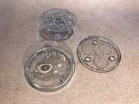 Three clear glass vintage cake plates displayed on carpet. One features a pedestal design, one includes a dome lid, and one is a flat plate with a clover motif.