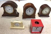 Five vintage clocks displayed on carpet: three wooden mantel clocks, one cream-colored desk clock, and one red Mickey Mouse clock. Visible wear and signs of age noted.