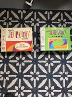 Two vintage Jeopardy! game boxes on patterned carpet - one Play Doh New Seventh Edition and one Milton Bradley New Jeopardy! Fifth Edition.