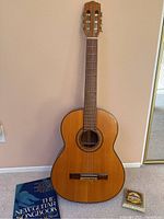 Full front view of acoustic classical guitar standing upright against wall with songbook and string pack on carpet next to it.