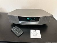 Front view of the Bose Wave Sound System showing digital clock display reading 12:11 and temperature 60°F. Includes remote control placed on the left.