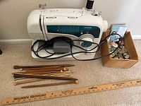 Full view of Brother sewing machine with grey foot pedal and attached power cable. Box next to machine contains sewing notions including thread spools and buttons. Several wooden knitting needles and crochet hooks laid in front with ruler for scale.