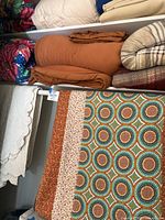 Overview of folded comforters and blankets including white comforter, rust-colored comforter, beige and tan blankets, and patterned comforter with geometric and floral design.
