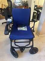 Front view of electric wheelchair showing blue mesh seat and backrest, joystick on right armrest, and included charger on seat.
