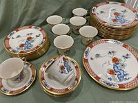 Overview of entire Lenox china set including stacked dinner plates, salad plates, cups, and saucers showing red lacquer border with blue and red floral design and gold trim.