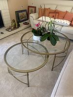 Photo showing all three nesting coffee tables arranged in a nested configuration on a beige carpet near a white sofa with orange pillows. Clear glass tops and light gold metal frames visible.