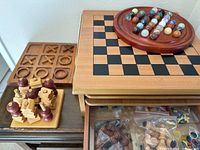 Photo showing chess and checkers board with carved chess pieces, a tic tac toe wooden game with X and O pieces, and partially visible stacked wooden game boards with drawers.