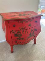 Front and side view of a vintage red dresser with two drawers and Asian style paintings on the surface and drawers. Shows signs of wear and patina.