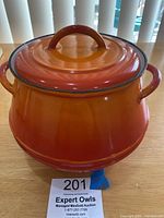Front view of vintage orange-red Descoware cast iron pot with lid on wooden table, showing shiny enamel finish and rounded shape with handles.