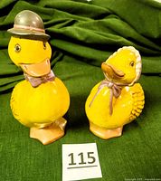 Front and side view of male and female ceramic duck figurines on green fabric background, showing details of hats, bows, and painted features.