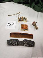 Antique and vintage jewelry accessories displayed together on white cloth, showing case, comb, pill box, locket with chain, and hand-shaped clip: overview shot