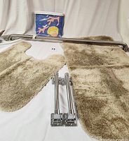 Photo showing tan contour and rectangular rugs, metal grab bars, and packaged seashore-themed shower curtain on white cloth background.