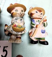 Photo of two gardening cat figurines, one holding a basket of flowers and the other a hoe, both wearing hats.