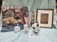 Lot shown on a chair with cat tapestry pillow, two ceramic white cats, framed ginger cat art, wooden cat figure, and a round white night light.