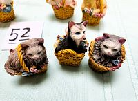 Three resin cat figurines in worn wicker baskets placed on fabric background, showing details of the cats and baskets