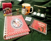 Overview photo showing all six items including cookbook, photo holder, figurine, golf balls in box, CD, and mug arranged on dark fabric background.