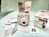 Full lot layout including white Fujifilm Instax Mini 9 camera, original box, two packs of instant film, instruction manuals, and camera strap on a light background.