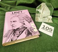 Photo of 1972 hardcover biography book 'Liszt' by Claude Rostand and clear glass piano sculpture on green velvet background.