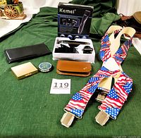 Full lot overview showing all items on green cloth background: Kemei hair clipper box and system, brown leather manicure case, square metal medicine case, round pill case, bifold wallet, and USA flag suspenders.