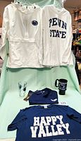 Photo showing white Penn State windbreaker and long-sleeve shirt hanging, navy glass and black mug in front, navy polo shirt and navy t-shirt folded below