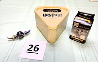 Photo of the lot including Harry Potter Trivial Pursuit game triangular plastic case, Dobby Funko POP keychain in box, and metal fidget orb spinner with wings design, on a beige fabric surface with numbered lot card.