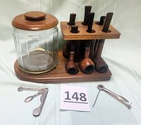 Full lot displayed including the clear glass humidor jar with wooden lid placed on a wooden base holding six pipes upright and two small metal pipe cleaning tools placed in front.