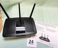 Top view showing the router with three antennas and ventilation grills, placed on a light-colored fabric surface with product sheet behind.