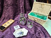 Photo shows the full set with deep purple fleece throw as backdrop. Clear glass hand-painted carafe and tumbler decorated with violets. Small white china dish with violet motif and wicker basket with lid. Box of Aldas soap with violet graphics.