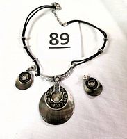 Top view of necklace and earrings laid flat, showing black double cords with silver rings, iridescent pendant with decorative silver and black top piece, and matching circular earrings with silver and black accents.